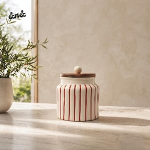Puede incluir: Un tarro cilíndrico de cerámica con base de color crema y rayas verticales rojas. El recipiente cuenta con una tapa plana de madera rematada con un pequeño pomo esférico de madera, que contrasta con el cuerpo de cerámica del tarro.
