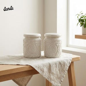 May include: A white ceramic mug with a black handle and the text "But First, Coffee" in a bold, black font. The mug is set against a blurred background, suggesting a cosy setting.