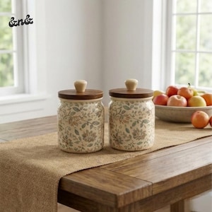 Op de afbeelding: Twee keramische potten met bloempatronen en houten deksels staan op een houten tafel. De potten hebben een crèmekleurige achtergrond met een ontwerp van groene, bruine en bruine bladeren en bloemen. Een fruitschaal staat op de achtergrond.