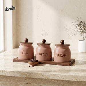 May include: Three dusty pink ceramic canisters with dark brown wooden lids and spherical knobs, labelled TEA, COFFEE, and SUGAR in black text. The canisters sit on a rectangular dark wood tray, accompanied by a small bowl of coffee beans.