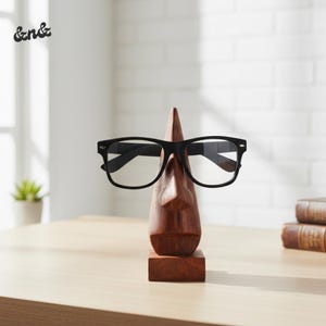 May include: A wooden eyeglass holder shaped like a face, with a nose and a square base. Black-framed eyeglasses rest on the nose. The holder is made of dark wood and sits on a light-colored wooden surface.
