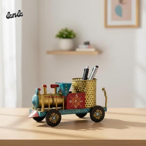 Puede incluir: Un organizador de escritorio decorativo con forma de tren en dorado, rojo y azul. El tren presenta un portalápices y ruedas decorativas. El organizador tiene las letras "&n&" en el lateral.