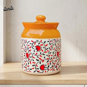 Op de afbeelding: Een keramische pot met deksel. De witte basis is versierd met een rood, zwart en wit bloemmotief. De bovenkant en het deksel zijn oranje. De pot staat op een houten plank.