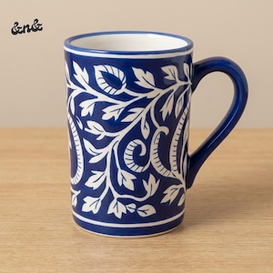 Puede incluir: Una taza de cerámica azul con un diseño floral blanco. La taza tiene un asa curva y un interior blanco. El exterior presenta un patrón repetido de hojas y vides estilizadas. La taza está sobre una superficie de madera marrón claro.
