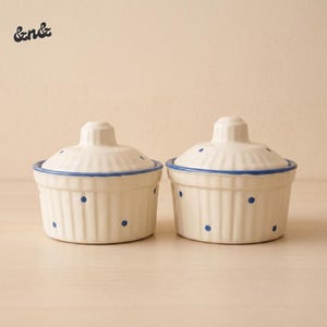 May include: Two white ceramic ramekins with lids. Each ramekin has a blue rim and blue polka dots. The lids have a small handle on top. The text "&nd&" is in the upper left corner. These are ideal for baking and serving.