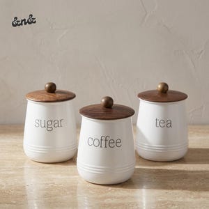 Op de afbeelding: Drie witte keramische voorraadpotten met donkerbruine houten deksels en bolvormige knoppen. Elke pot heeft een geribbelde basis en zwarte tekstlabels met de woorden sugar, coffee en tea, opgesteld in een rij op een ondergrond.