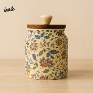 Peut inclure: Un pot en céramique avec un couvercle en bois et un bouton clair. Le pot est décoré d'un motif floral dans les tons de bleu, orange et vert sur un fond crème. Le pot est posé sur une surface en bois.