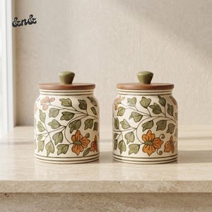 Può includere: Due barattoli cilindrici in ceramica con coperchi in legno, caratterizzati da un motivo botanico dipinto a mano con fiori arancioni e foglie verdi su sfondo crema. Il testo "&n&" appare nell'angolo in alto a sinistra dell'immagine.