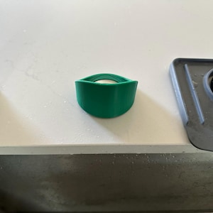 May include: A green, curved soap dish with a white bar of soap inside. The dish is made of a flexible material and sits on a white countertop near a stainless steel faucet and a dark gray sink.