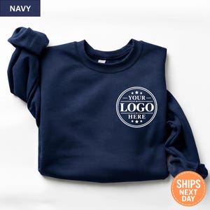 May include: A navy blue crewneck sweatshirt featuring a white circular graphic on the left chest. The graphic contains the text "YOUR LOGO HERE" surrounded by decorative lines and stars. The sweatshirt has long sleeves and a ribbed neckline.