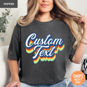 May include: A charcoal gray short-sleeve t-shirt featuring the words Custom Text in a white script font. The text is layered over a multi-colored retro-style drop shadow design in shades of blue, orange, yellow, and light green.
