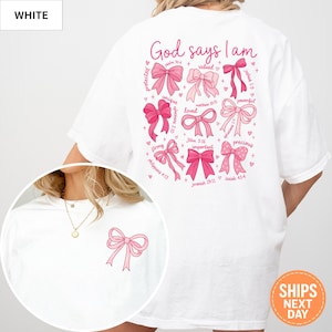 May include: White t-shirt with a pink bow graphic on the front and a larger design on the back. The back design features pink bows with the words "God says I am" and inspirational words like "loved" and "precious".