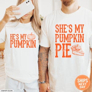 May include: Two off-white t-shirts featuring orange text and graphics. The left shirt displays the text HE'S MY PUMPKIN with a pumpkin illustration. The right shirt displays the text SHE'S MY PUMPKIN PIE with a slice of pumpkin pie illustration.