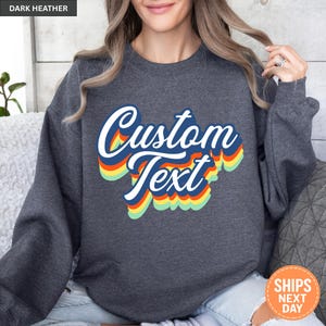 May include: A dark heather gray crewneck sweatshirt featuring the words Custom Text in a bold, white script font. The text is layered with a retro-inspired, multi-colored drop shadow effect in shades of blue, orange, yellow, and light green.