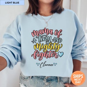 May include: Light blue sweatshirt with the text "Mama of a Tiny and Mighty Fighter" in pink, yellow, and blue. The name "Clarence" is below the text, with star accents. The sweatshirt is a solid light blue color.