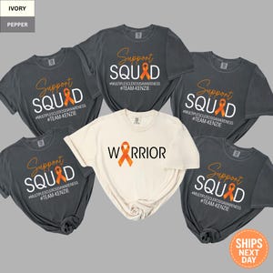 May include: A collection of t-shirts in ivory and pepper colors. The shirts feature the words "Support SQUAD" and "WARRIOR" with an orange ribbon, promoting multiple sclerosis awareness. The text "#TEAM-KENZIE" is also included.