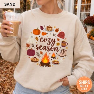 May include: A sand-colored crewneck sweatshirt featuring a central graphic with the text cozy season surrounded by illustrations of a pumpkin, ghost, campfire, pie, boots, mug, and autumn leaves in warm orange, brown, and purple tones.