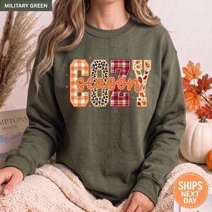 May include: A military green long-sleeve sweatshirt featuring the text COZY season in a block-letter design. The letters are filled with orange plaid, leopard print, red plaid, and autumn leaf patterns, accented by small orange star shapes.