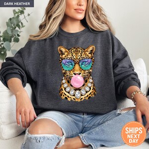 May include: A dark heather gray crewneck sweatshirt featuring a graphic of a leopard wearing colorful patterned sunglasses and a large rhinestone necklace while blowing a pink bubblegum bubble. The leopard has yellow and black spots.