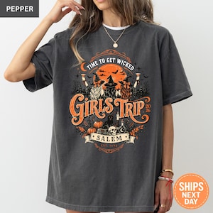 May include: A charcoal gray t-shirt featuring a graphic print with three witches in hats, a black cat, pumpkins, a skull, and the text "TIME TO GET WICKED", "GIRLS TRIP 2026", and "SALEM EST. 1692" set against a large orange moon and bats.