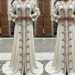 Puede incluir: Vestido crema de manga larga con bordados florales en tonos pastel de rosa, azul y morado. El vestido presenta una capucha y una línea vertical de cuentas perladas en la parte delantera. Las mangas y los paneles frontales están adornados con motivos florales.