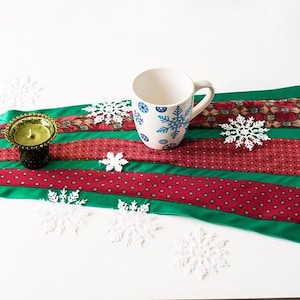 Pode incluir: Um caminho de mesa de Natal festivo feito de gravatas reaproveitadas. O caminho de mesa apresenta seções verdes e vermelhas alternadas com detalhes de flocos de neve. Uma caneca branca com flocos de neve azuis e um castiçal com uma vela acesa estão em cima.