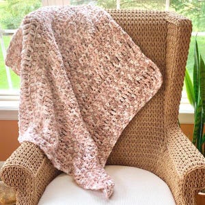 May include: A soft, chunky knit blanket in shades of pink and cream is draped over a wicker chair. The blanket's texture is visible, with a thick, plush appearance. The chair has a woven design and a white cushion.