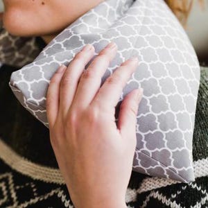 May include: A gray and white patterned neck wrap is held around a person's neck. The wrap has a geometric design and is made of soft fabric. The person is wearing a green and black sweater.