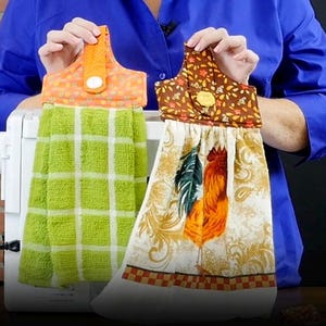 May include: Two decorative kitchen towels. One is green with a white grid pattern, and the other features a rooster design with orange, green, and brown accents. Both towels have a loop for hanging and are held up by a person.