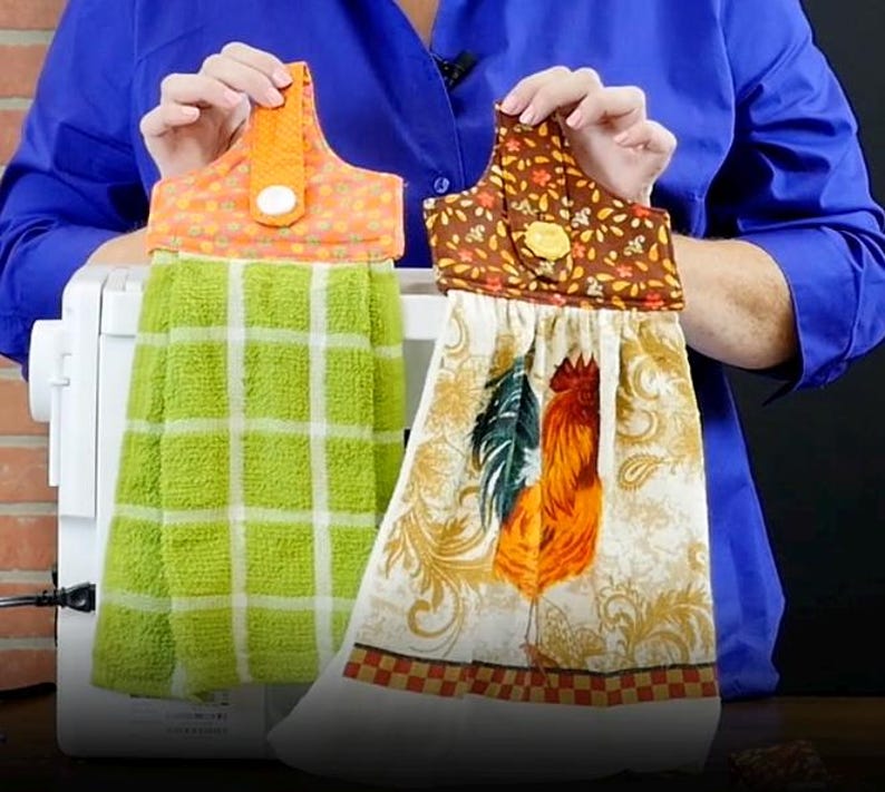 May include: Two decorative kitchen towels. One is green with a white grid pattern, and the other features a rooster design with orange, green, and brown accents. Both towels have a loop for hanging and are held up by a person.