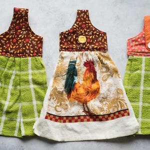May include: Three decorative kitchen hand towels. The towels feature a loop for hanging and a towel portion. One towel has a rooster design, and two have a green and white plaid pattern. The tops have floral and polka dot patterns.