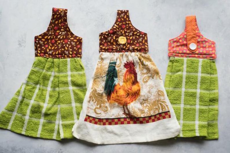 May include: Three decorative kitchen hand towels. The towels feature a loop for hanging and a towel portion. One towel has a rooster design, and two have a green and white plaid pattern. The tops have floral and polka dot patterns.