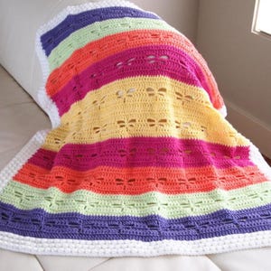 May include: A colorful, handmade crochet blanket with horizontal stripes in shades of purple, green, orange, pink, and yellow. The blanket features a white border and dragonfly cutouts, creating a textured and vibrant design. The blanket is draped over a white surface.