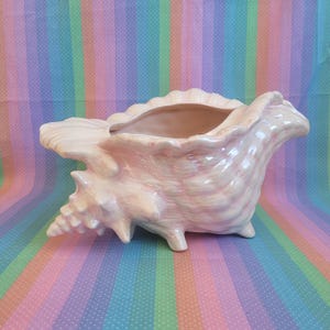 May include: A glossy, pale pink ceramic planter shaped like a large conch shell. The surface features a smooth, iridescent glaze with subtle variations in colour, highlighting the natural ridges, spiral details, and pointed protrusions of the shell.