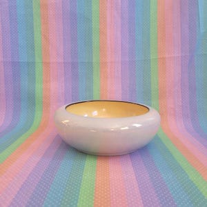 May include: A low, round ceramic bowl with a smooth, iridescent white exterior finish and a contrasting pale yellow interior. The bowl features a simple, wide-rimmed design with a dark, thin line accenting the top edge of the opening.