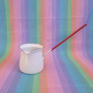 May include: A white enamel milk pot with a long, thin handle featuring a red accent. The pot has a rounded body, a wide rim, and a small pouring spout. The metal construction is finished with a smooth, glossy white coating throughout the exterior.