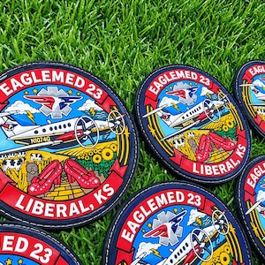 Könnte beinhalten: Runde gestickte Aufnäher mit einem weißen Flugzeug mit roten Akzenten, einem blauen Himmel mit Blitzen, einer gelben Backsteinstraße, Sonnenblumen, einem kleinen Hund und roten Schuhen. Der Text lautet EAGLEMED 23 und LIBERAL, KS.