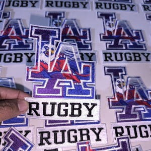 Puede incluir: Un parche bordado rectangular que presenta las letras LA en una fuente estilizada con un gráfico de águila azul y roja integrado en el diseño. Debajo de las letras, una pancarta rectangular blanca muestra la palabra RUGBY en texto negro.