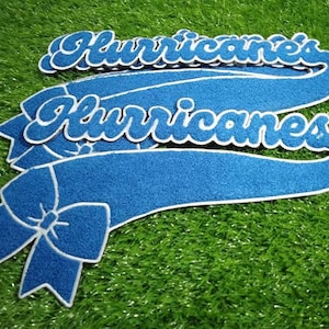 Puede incluir: Bandera de fieltro azul con la palabra "Hurricanes" en escritura blanca. La bandera tiene un detalle de lazo y un borde blanco. Los art&iacute;culos est&aacute;n sobre una superficie verde texturizada.