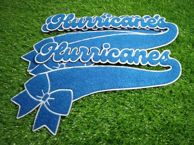 Puede incluir: Bandera de fieltro azul con la palabra "Hurricanes" en escritura blanca. La bandera tiene un detalle de lazo y un borde blanco. Los art&iacute;culos est&aacute;n sobre una superficie verde texturizada.
