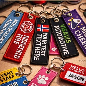 May include: A collection of personalized keychains in various colors, each with custom text and designs. The keychains feature embroidered text, logos, and a metal ring for attachment. The image includes keychains with the text "YOUR TEXT HERE", "SARAH", and "JASON".