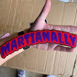 Puede incluir: Un parche bordado rojo y morado con la palabra "MARTIANALLY" en un dise&ntilde;o curvo. El parche tiene un borde negro y se muestra en el brazo de una persona.