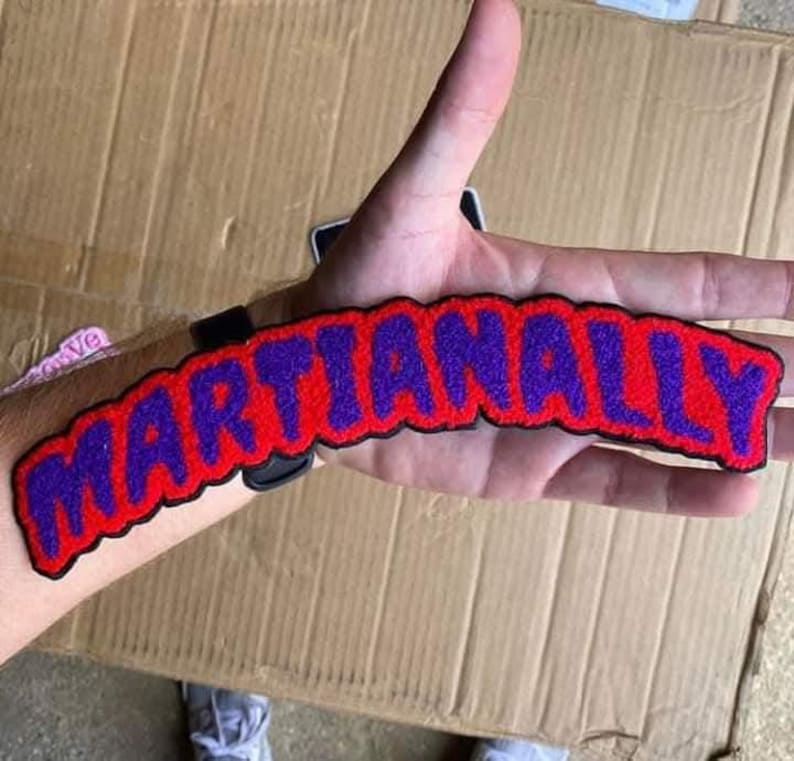 Puede incluir: Un parche bordado rojo y morado con la palabra "MARTIANALLY" en un dise&ntilde;o curvo. El parche tiene un borde negro y se muestra en el brazo de una persona.