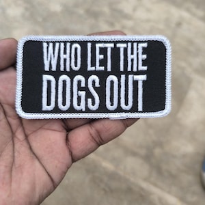 May include: A rectangular embroidered patch with a black background and a white border. The text WHO LET THE DOGS OUT is stitched in white capital letters across the center of the patch. The patch features a clean, stitched edge finish.