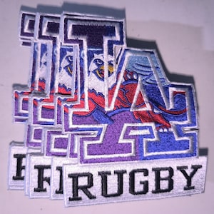 May include: A stack of embroidered patches featuring a large purple and blue letter A with a white eagle graphic inside. Below the letter, a white rectangular banner contains the black text RUGBY. The patches have a white border.