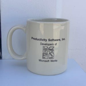 May include: A cream-colored ceramic mug featuring black printed text that reads Productivity Software, Inc. Developers of WP, DB, SS, COM and Microsoft Works, accompanied by four small square icons representing different software applications.