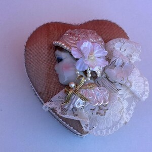 May include: A heart-shaped brooch featuring a porcelain face profile wearing a textured pink hat with a pearl trim. The figure is adorned with a light pink fabric flower, a gold ribbon bow, and white lace accents layered over a brown fabric base.