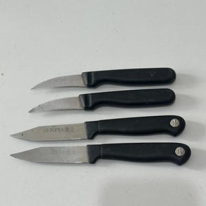 May include: Four small kitchen knives with black handles and silver blades. Two knives have curved blades, and two have straight blades. The brand "DOMUS" is visible on two of the knives. The knives are arranged on a white surface.