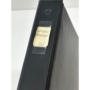 Può includere: Un raccoglitore nero con un'etichetta che recita "HP9820 PROGRAMS" in testo nero. L'etichetta è fissata con nastro adesivo trasparente. Il raccoglitore è leggermente angolato, mostrando il dorso e parte della copertina.