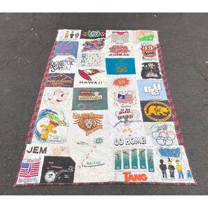 May include: A rectangular memory quilt constructed from various graphic t-shirt panels featuring diverse prints, including cartoon characters, university logos, sports themes, and text such as 'JEM', 'GO HOME', and 'DIABLO DIVERS', finished with a red plaid border.